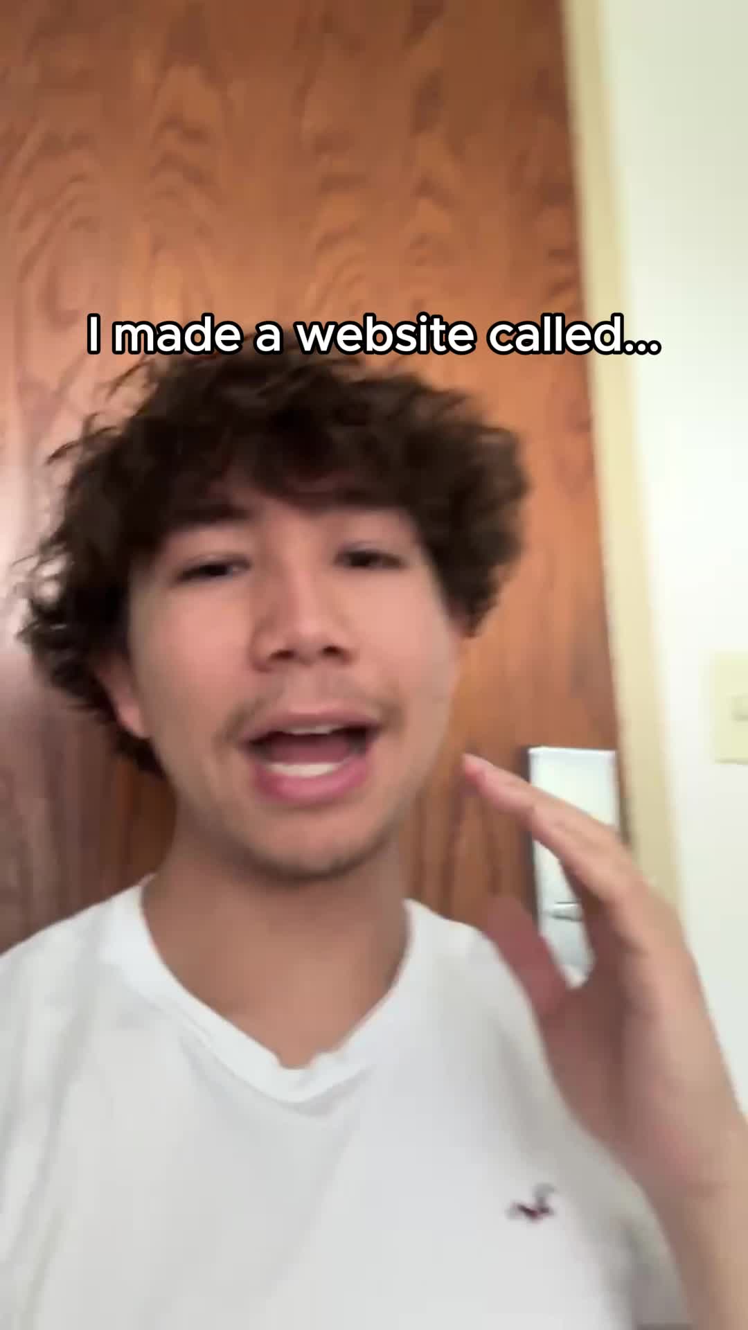 A young man with short, dark curly hair is centered in the frame, facing the camera and speaking. He wears a white t-shirt. The background is a light-colored wall and a wooden door. Text overlays the top, reading "I made a website called..."