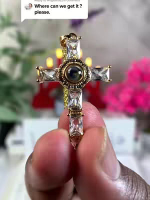 A close-up shot of a gold cross pendant encrusted with sparkling clear gemstones. The cross is held between two fingers against an out-of-focus background of soft greenery and what appear to be candle-like lights. A comment bubble is visible in the upper-left corner with overlaid text.