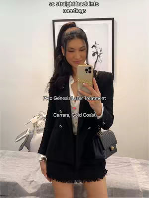 A woman takes a mirror selfie while holding her phone up.  She has long, dark hair pulled back in a high ponytail, wearing a black blazer and skirt, and a black purse. The background is a mix of white wall and a framed picture, indicating a clinic setting.