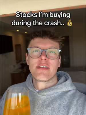The cover image shows a man in a gray hoodie holding a clear glass filled with an orange liquid, possibly a mimosa. The man is centered in the frame, and has glasses and short hair. Above him in bold, white text reads "Stocks I'm buying during the crash.." with a money bag emoji next to it. The background is softly blurred, showing a room.