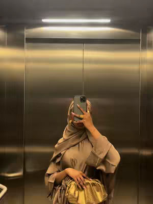 A woman in a hijab takes a mirror selfie in an elevator. She is wearing a light brown abaya. The elevator walls are a metallic color, and a bright light is seen above.
