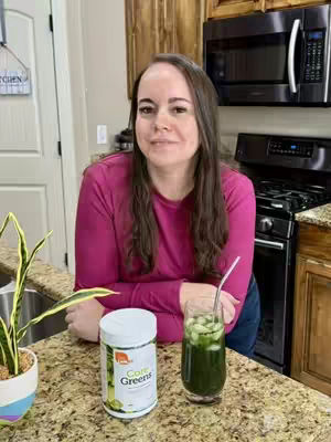 A woman in a pink long-sleeved shirt stands in a kitchen, leaning against a countertop. On the counter, a container of 'Core Greens' and a glass of green beverage with a straw are visible. The background shows kitchen cabinets and appliances, including a microwave.