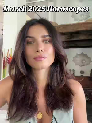 A woman with long brown hair looks directly at the camera. She wears a light-colored top and a gold necklace. The background is slightly out of focus, showing a wooden structure and some plants. Text overlay at the top says 'March 2025 Horoscopes.'