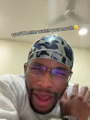 A selfie-style video featuring a man with a durag, glasses, and an open mouth, suggesting he's about to speak. The background is a blurry indoor setting.