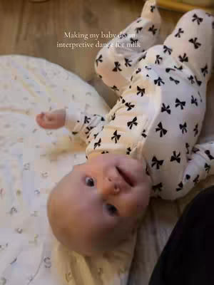 A baby is laying on its back on a white patterned blanket, angled diagonally across the frame. The baby is dressed in a black bow-tie patterned onesie, and is looking at the camera with its head turned towards the right side of the image.
