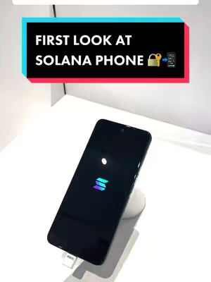 A black smartphone on a white stand is displayed on a white surface. The phone screen shows a colorful logo. Above the phone is a rectangular overlay with the text 'FIRST LOOK AT SOLANA PHONE'.