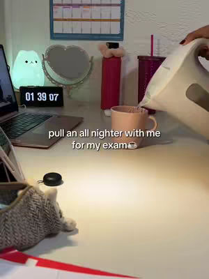 A person is shown preparing a hot drink while studying. A cup is being filled with hot water from a kettle. In the background, a laptop shows a screen displaying what seems to be the study material. The setting includes study room with items arranged on a desk. There's a light-up decorative item, a mirror, stationary items, and a calendar.