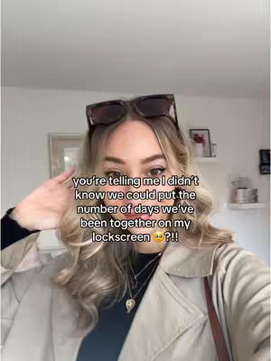 A woman with long blonde hair, wearing a beige trench coat, and sunglasses on her head, is looking at the camera. Text overlayed on the image asks if she knew she could put the number of days she's been with her partner on her lockscreen.
