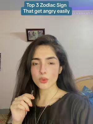 A woman with long dark hair is centered in the frame, holding a small black object near her lips. She is wearing a black top and a silver necklace. Above her head, a teal banner reads, "Top 3 Zodiac Sign That get angry easily." The background is a light-colored wall with a framed picture. The overall composition suggests a personal, informational video.