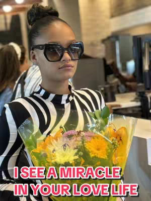 A woman with dark hair in a bun, wearing large black sunglasses and a black and white patterned top, holds a bouquet of flowers. Text overlayed on the image reads 'I SEE A MIRACLE IN YOUR LOVE LIFE'. The background appears to be a public space with softly focused elements and natural light.