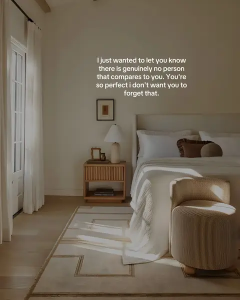 A well-lit interior shot of a bedroom or living space. The room features a bed with white linens, a small side table with a lamp, and a patterned rug. Natural light streams in from a window, illuminating the scene. The overall ambiance is peaceful and serene.
