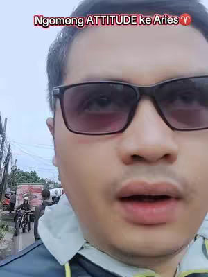 A man wearing black-framed sunglasses stands outdoors, with a blurry background of a street scene including vehicles and other people. The man's face fills most of the frame, with a text overlay at the top that has a red background.