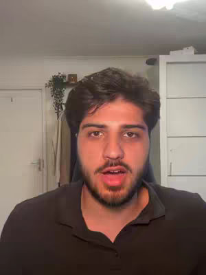 A frontal view of a young man with dark hair and stubble, speaking directly to the camera. He wears a black shirt and is in a well-lit room. The background includes a white cabinet and a hint of decor, suggesting a home or casual setting.