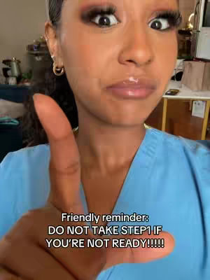 A close-up selfie features a light-skinned woman with dark hair styled back, wearing makeup that includes bold eye shadow and glossy lips. She is pointing a finger towards the camera, with the focus on her face and hand. The background is slightly blurred, but hints at an indoor setting.