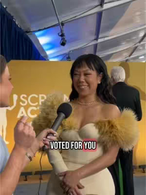 A woman stands in front of a yellow background with a microphone in front of her while another person is holding the microphone, and there is a text overlay.