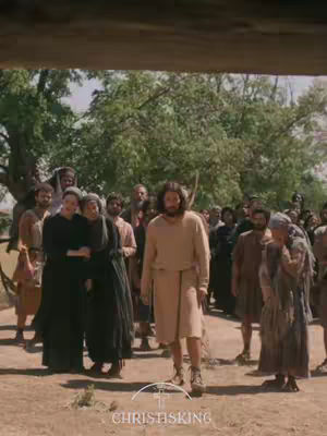 A scene from a movie set outdoors with several people dressed in biblical attire. Jesus, wearing a light-colored robe, stands in the center with a crowd surrounding him.