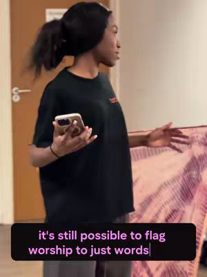 A woman in a black shirt holds a phone and a fabric, standing inside a room. Light walls and a partly visible door contribute to the indoor setting.