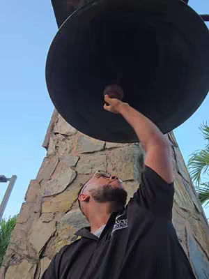 A man is reaching up to touch the clapper of a large, dark bell. The bell is suspended from a stone structure. The man is wearing glasses and a black collared shirt, looking up with a serious expression. The background shows a clear blue sky and a few palm trees peeking into frame.