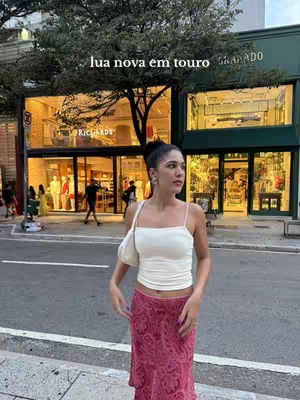 A woman stands on a city street in front of a colorful storefront. She wears a white top and a long, pink skirt. The image is bright and the composition is balanced.