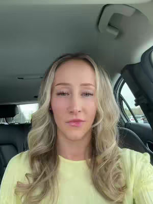 A young woman inside a car looking at the camera. She has long blonde hair and is wearing a yellow shirt. The background shows the interior of the car and the street outside.