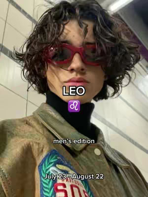 A person with curly, dark hair and red sunglasses is in the center of the frame. The person wears a black turtleneck and a brown and green jacket. Text overlays include 'LEO' with the Leo zodiac symbol, 'men's edition', and 'July 23 - August 22'. The background is a blurry, light-colored wall.