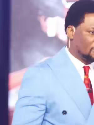 The image shows a man in a light blue suit jacket over a white dress shirt and a red tie. He appears to be on a stage or in front of a blurred background. The image is cut off on the left side, showing only a portion of the man's face and shoulder.