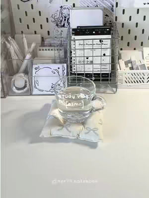 A well-lit, top-down shot of a clean, organized desk setup. A glass cup with water sits on a small bow-adorned cushion in the foreground. Behind it, various desk organizers and supplies are partially visible. The background features more storage solutions and a pegboard. The color palette is soft and neutral.