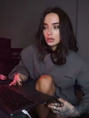 A medium shot of a woman sitting in a dimly lit room, with a laptop on her lap. She is wearing a gray hoodie and several rings. The background is dark and out of focus.