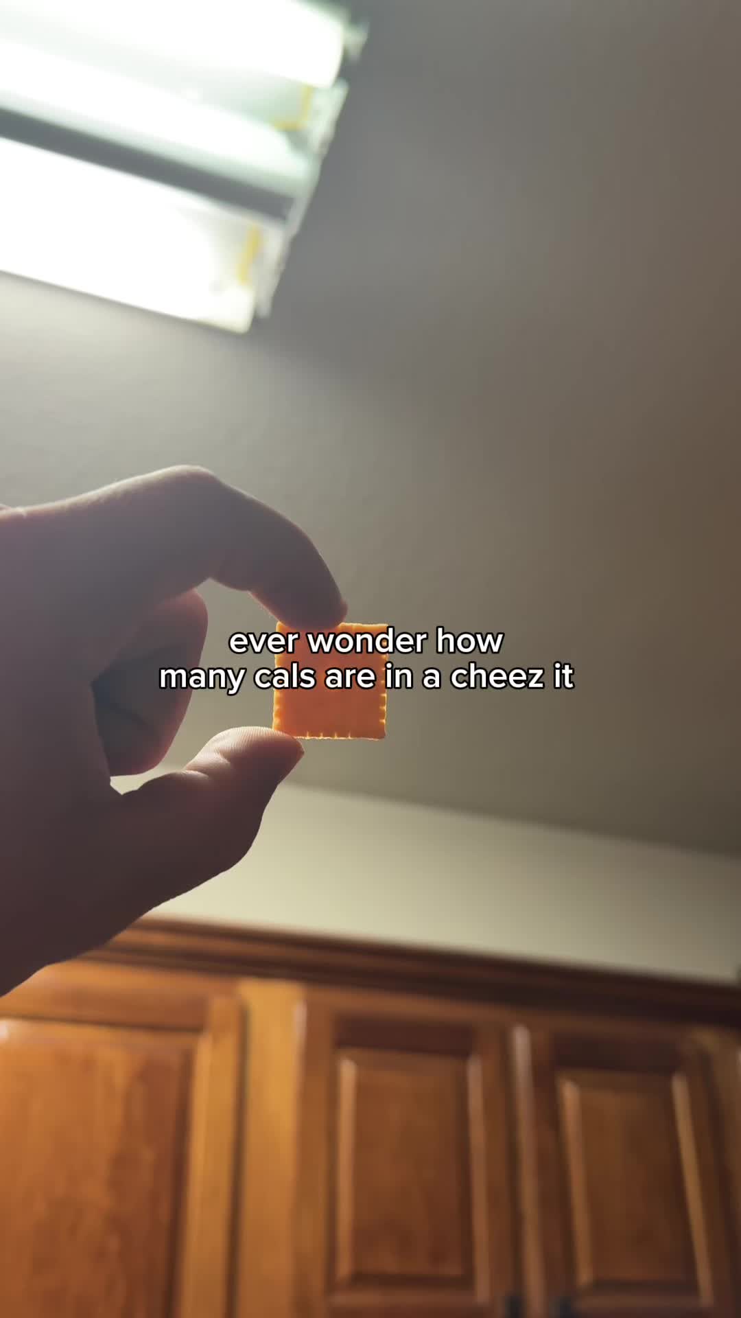 A hand holding a single Cheez-It cracker with the image focusing on the Cheez-It. In the background, a light fixture and wooden cabinets are visible.