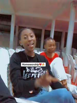 A woman with dark hair, wearing a black long-sleeved shirt and blue jeans, is the primary focus. She is seated, gesturing expressively, and has a focused expression. A blurred background includes another person in a red shirt and white walls, suggesting an indoor setting. A text overlay reads "Homeworship" centered in a white square, and another text is visible at the bottom, potentially a caption or additional message.