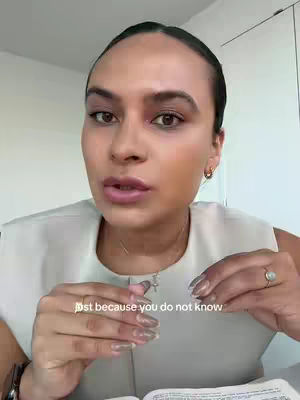 A person with dark hair and brown eyes is centered in the frame, with a light neutral-colored top. The person is speaking, with text overlayed. The neutral background suggests a well-lit indoor setting.