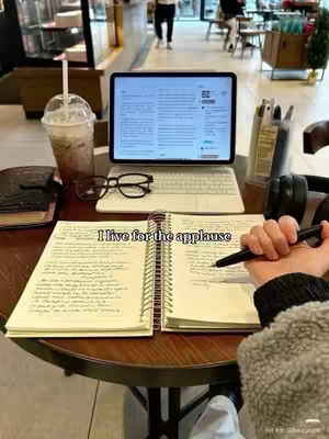 A person is working on their laptop and writing in a notebook on a round wooden table, with a drink, glasses and headphones. The text overlay reads: 'I live for the applause'. The background is blurred out, but appears to be a cafe setting, with other people present.