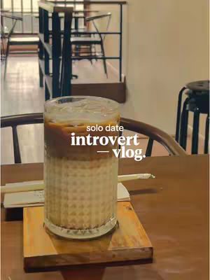 An overhead shot features a tall glass of iced coffee sitting on a wooden coaster. The beverage is a mix of layered textures: a light-colored bottom, then a creamy middle, and coffee on top. The glass is placed on a square wooden coaster. This is all set on a wooden table. In the background, a blurred interior setting with structural elements.