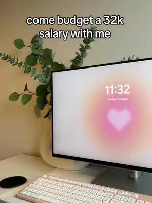 A well-lit workspace featuring a computer with a soft, pink and orange gradient background and a heart-shaped design. A green leafy plant sits in a white vase next to the monitor. A white keyboard and a black mouse pad are visible.