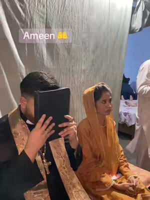 The image shows a man in religious attire holding a book in front of his face, with a woman beside him wearing a head covering. A text overlay reads 'Ameen' and the overall setting appears to be a solemn event.