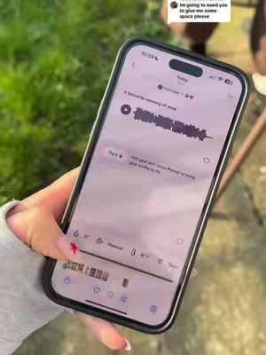 A person's hand holding a smartphone with a black case, partially obscuring an app screen. The app screen displays text and interactive elements. The background shows green grass.
