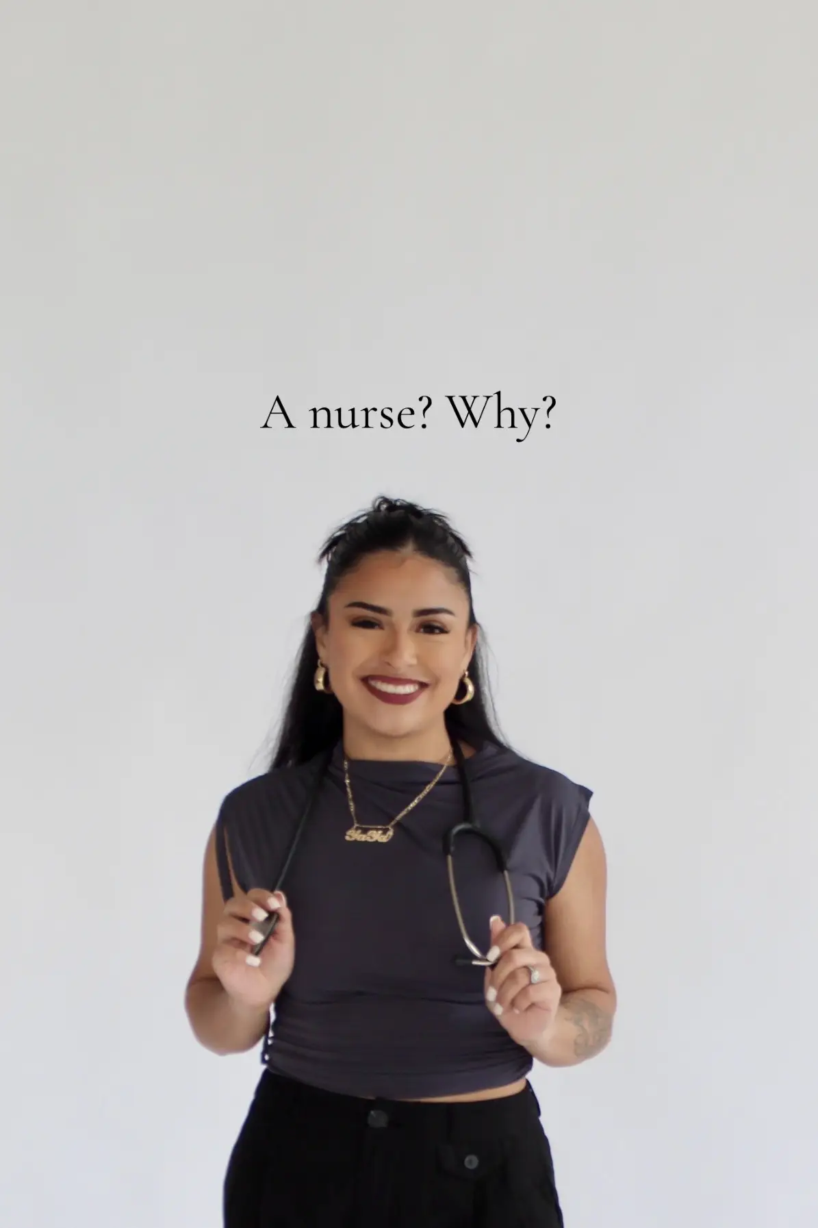 A woman with dark hair smiles at the camera. She wears a dark gray top and black pants. She's holding a stethoscope, indicating she may be a healthcare worker. Text overlays the image: "A nurse? Why?"