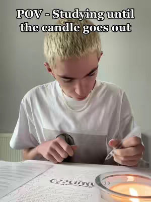 A young man with short blond hair is sitting at a table, focused on studying. He is wearing a white t-shirt and has a delicate necklace. On the table in front of him are open books and a burning candle in a glass bowl; the text overlay reads "POV - Studying until the candle goes out". The background is a plain wall with a radiator.