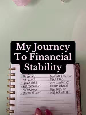 The cover image shows a spiral notebook open to a page of handwritten notes. A black rectangular banner towards the top reads "My Journey To Financial Stability". The notebook displays a list of financial issues and aspirations, written in black ink. In the background are the edges of a table and part of an open planner showing a pink crystal.