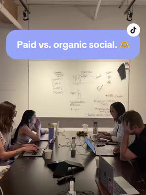 A group of five people around a large table, presumably in an office setting. They are all facing the same direction with their backs to the camera, working on laptops. A whiteboard is visible, with some writing on it. A text box overlay at the top of the image reads Paid vs. organic social.