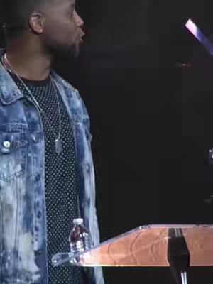 The image shows a man speaking at a lectern, with a microphone visible. He wears a denim jacket over a patterned shirt and a silver pendant. A partially visible bottle of water sits on the lectern. The setting is dark, with bright lights.