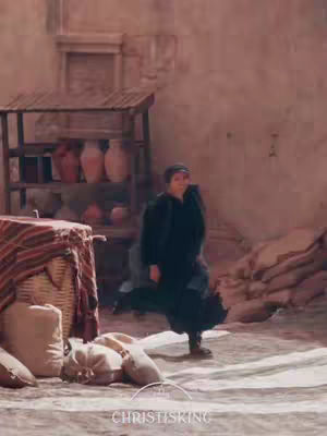 The image shows a woman in a long, dark dress and head covering, walking on a cobblestone path. She is positioned in front of a rustic building with arches, shelves holding pottery, and sacks on the ground, with a light-brown and muted color palette. The scene seems to be from a depiction of a historical setting, possibly religious, with a subtle watermark at the image's base.