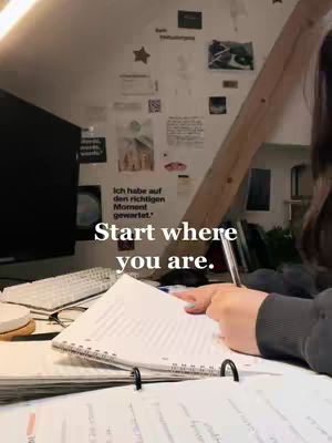 A slightly high-angle shot of a workspace reveals a woman's arm and hand in the foreground, focused on writing in a spiral notebook. The background shows a cozy study room with items attached to the wall along with the light from the scene.