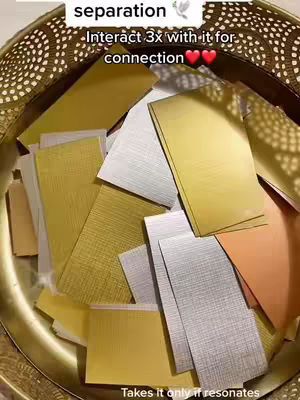 The cover image shows a close-up shot of a gold-colored decorative bowl filled with assorted rectangles. The rectangles vary in color and texture, including gold, silver, and peach, and potentially represent cards or slips of paper. The lighting is diffused, casting soft shadows within the bowl, and the background suggests a textured surface.