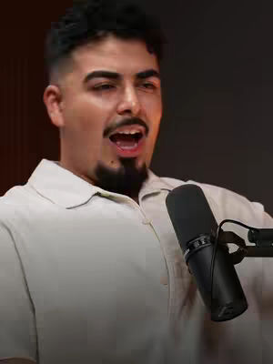 A man with short, dark hair, a well-groomed beard, and an open mouth, as if in mid-speech. He wears a collared shirt and holds a black microphone close to his face. The background is a blurred, dark shade.