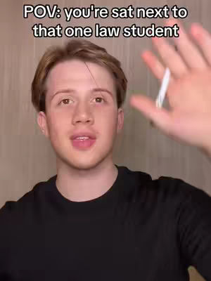 A young man with light skin is centered in the frame with text overlayed at the top. He is wearing a black shirt and has short, light-brown hair. He appears to be speaking, with his mouth open and his right hand raised, as if in a gesture to the side. The background is a plain, neutral color.