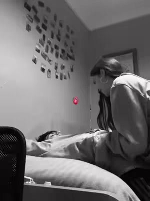 A black and white image showing a bedroom scene with a woman leaning over a person lying in bed. A collection of photos is on the wall above the bed, and a red heart graphic floats in the air.