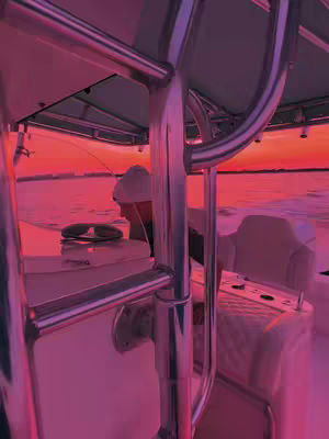 The image shows a man on a boat at sunset. He is wearing a white hat and is slightly hunched over. Visible on the boat are a white surface, metallic railings, and some fishing gear.