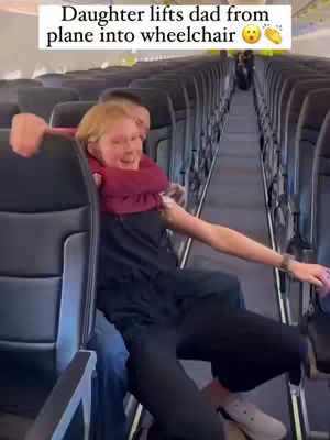 A blonde woman carrying a man, presumably her father, on a plane. She is smiling and appears to be helping him from his seat to a wheelchair. The background shows the interior of an airplane with seats and a central aisle.