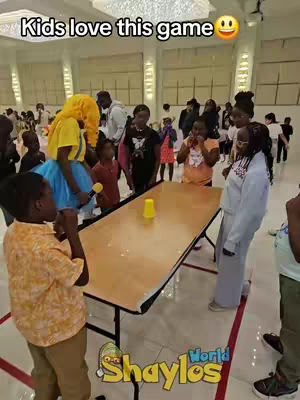 A vibrant scene from a children's party, with a large crowd of kids and adults gathered in a brightly lit hall. A long table takes center stage, with a single yellow cup on it. A person dressed in a yellow costume with a microphone appears to be hosting a game. The floor has a red and white pattern, and the image is framed with logos.